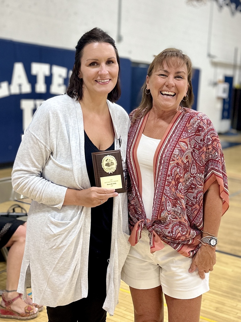 Two women stand together smiling; one holds a plaque award while the other has their arm around her.