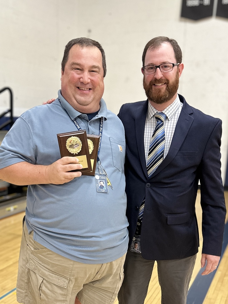 Two men stand together smiling; one holds a plaque award while the other has his arm around him.