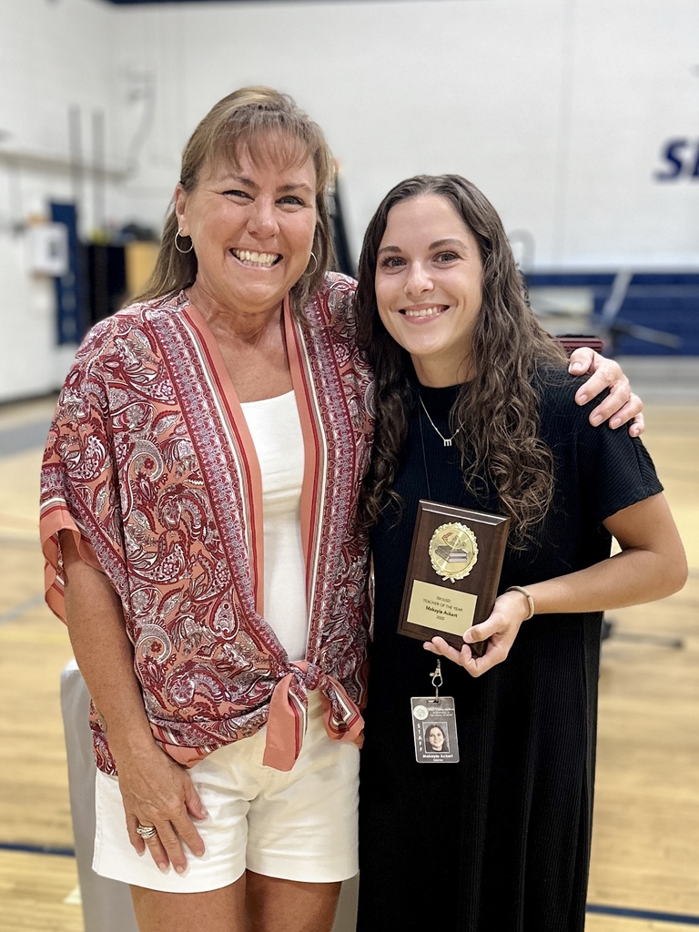 Two women stand together smiling; one holds a plaque award while the other has their arm around her.