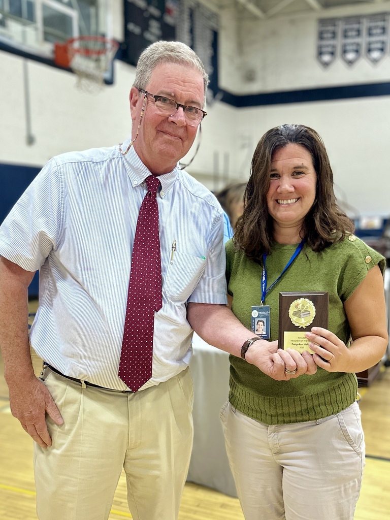 A man and woman smile while holding a plaque award together inside a gymnasium.