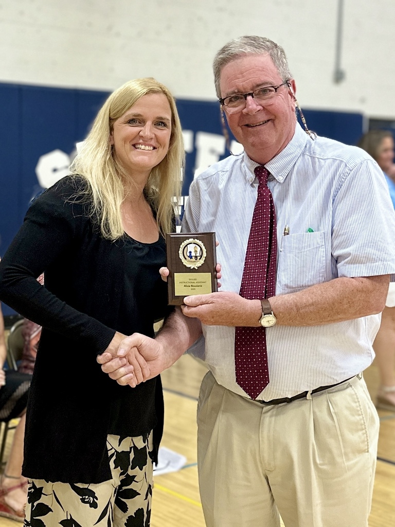A man and woman smile while holding a plaque award together inside a gymnasium.