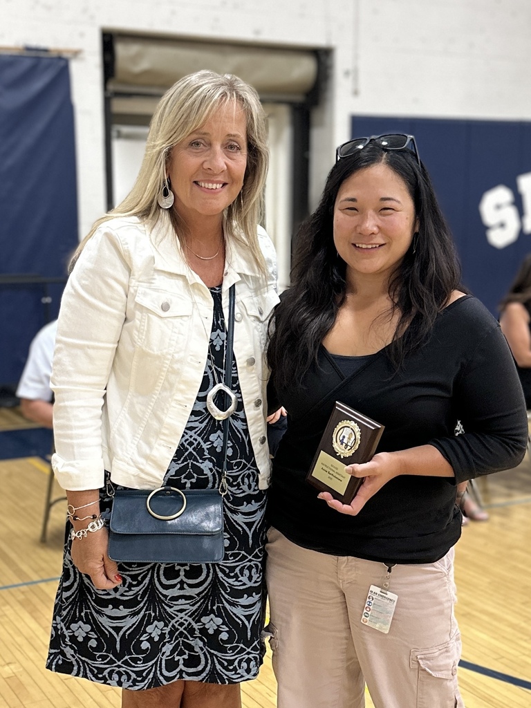Two women stand together smiling; one holds a plaque and both have their arms around each other.