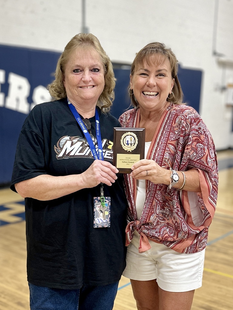 Two women stand together smiling holding a plaque award in a gymnasium.