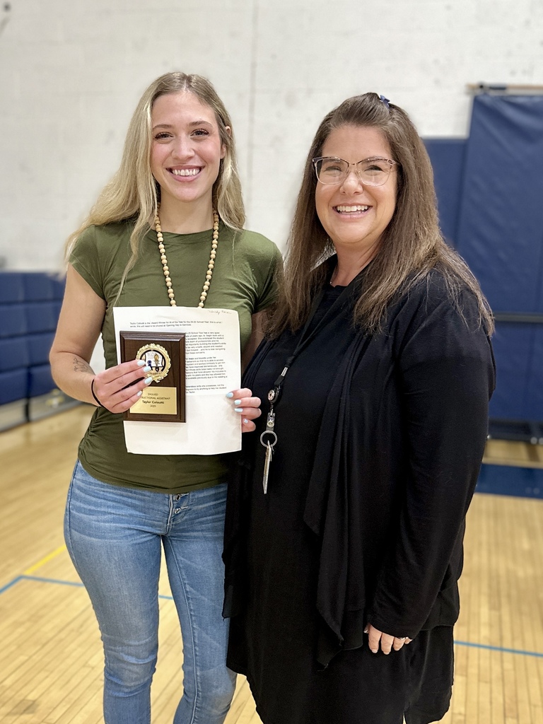 Two women stand together smiling; one holds a plaque while the other has their arm around her.