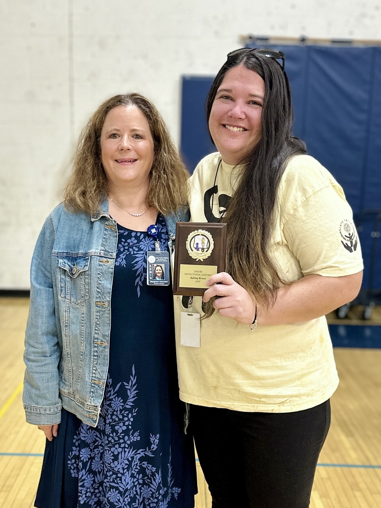 Two women stand together smiling; one holds a plaque and both have their arms around each other.