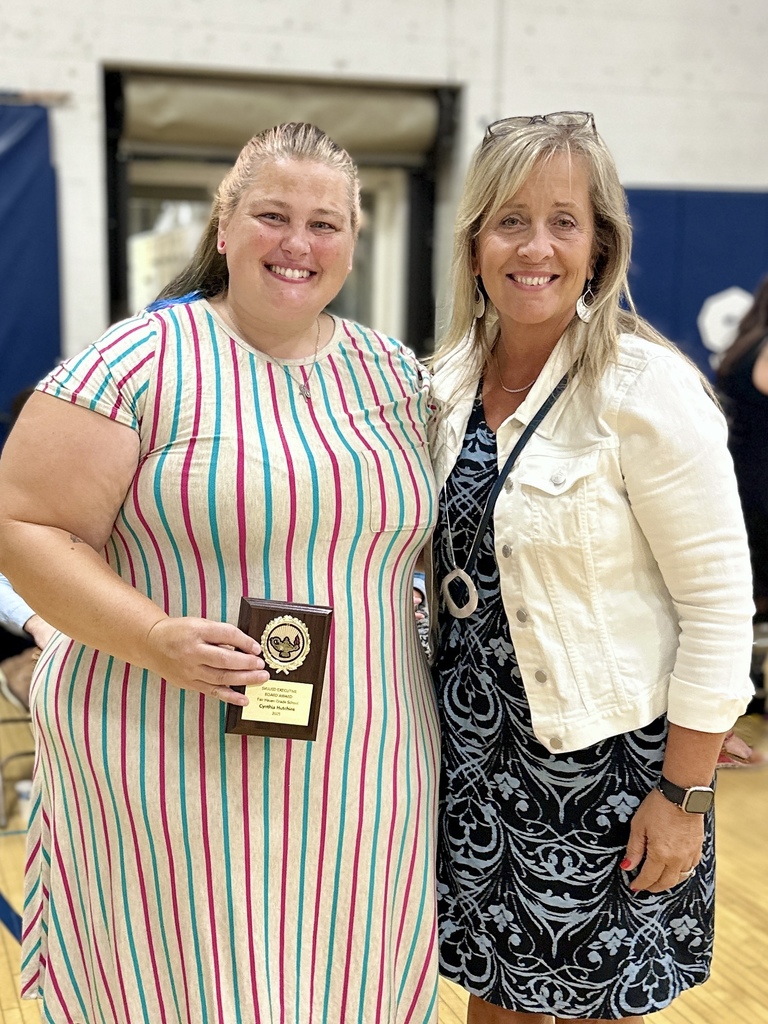 Two women stand together smiling; one holds a plaque award while the other has their arm around her.