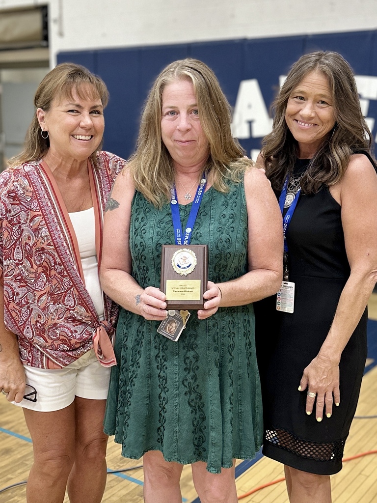 Three women smiling and standing together. The one in the middle holds an award plaque, while the other two women have their arms around the middle woman's back.