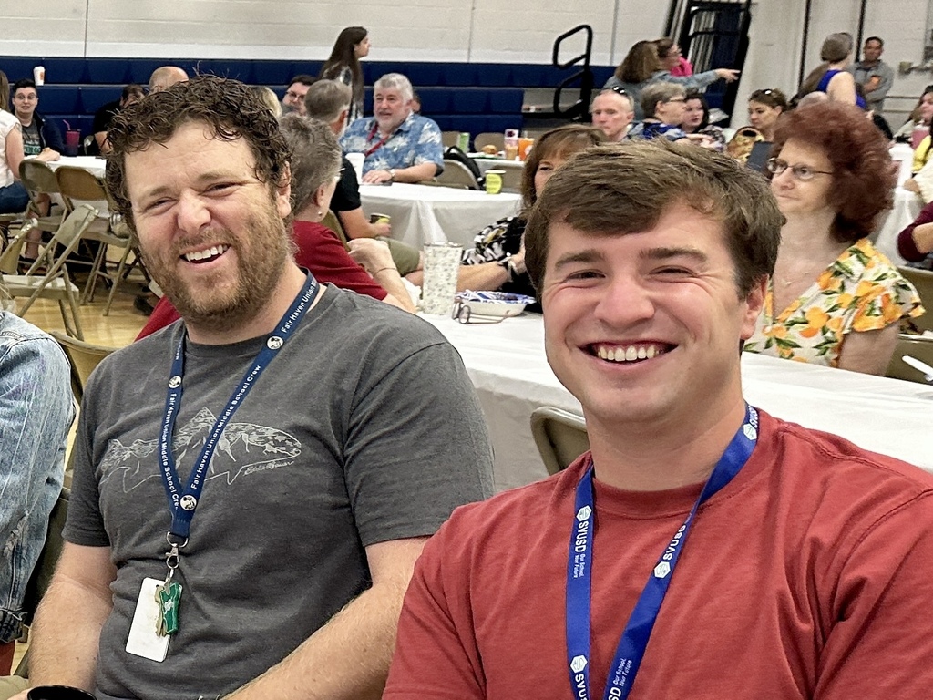 Two men wearing lanyards sit together at a table in a gymnasium, smiling at the camera.