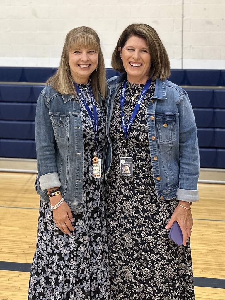 Two women smile and stand side by side in a gymnasium, both wearing denim jackets over long floral dresses with Slate Valley lanyards around their necks.