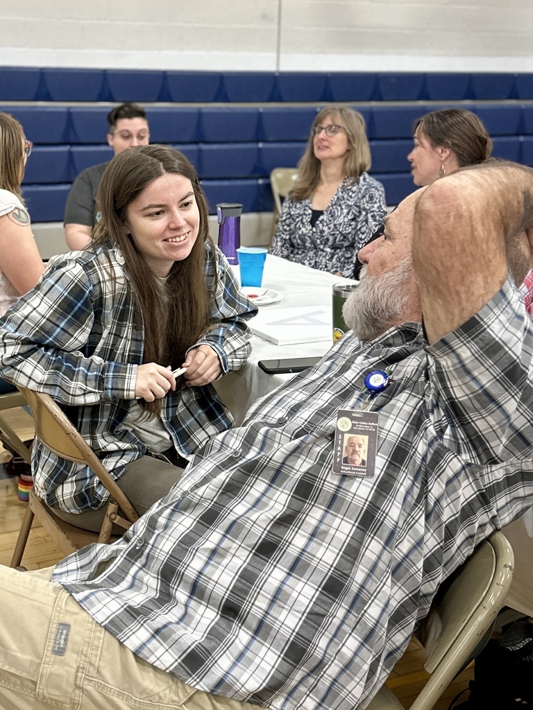A young woman and an older man sit at a table in a gymnasium, smiling and talking with others seated around them.