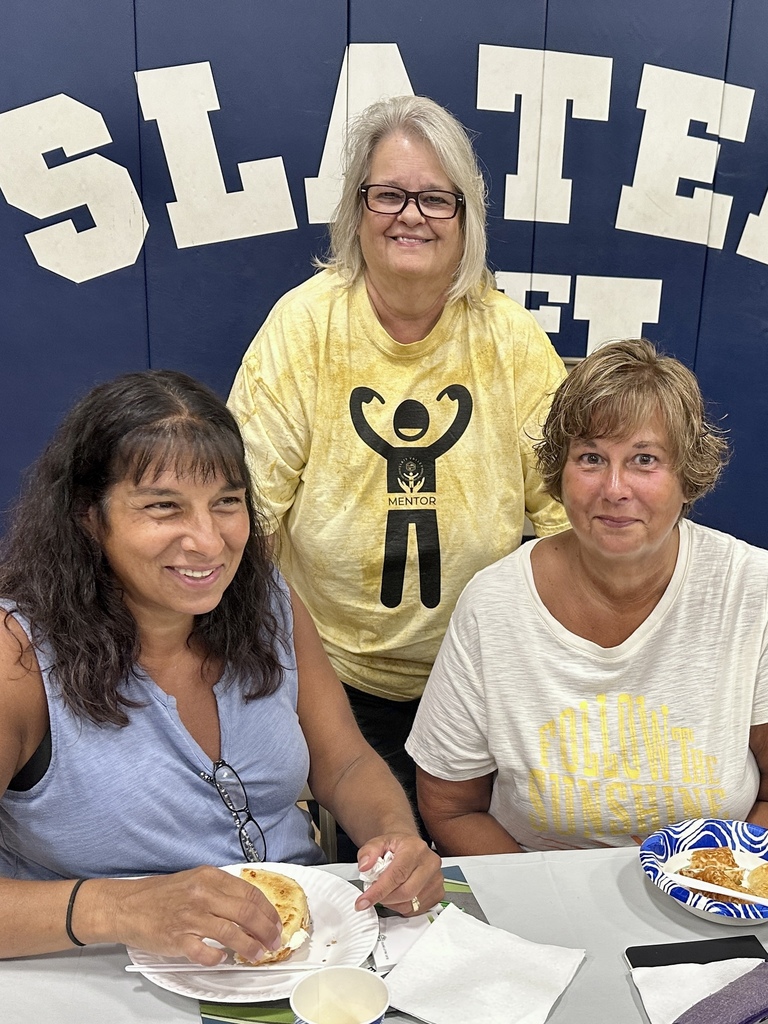 Three women smile while sitting together at a table in a gymnasium. Two are seated with plates of food, and one stands behind them wearing a yellow shirt with the word "Mentor." The background shows blue gym wall padding with large white letters.