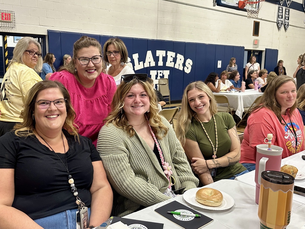 A group of five women sit and smile at a table during a school event, with notebooks, food, and drinks in front of them, while more people gather in the background.