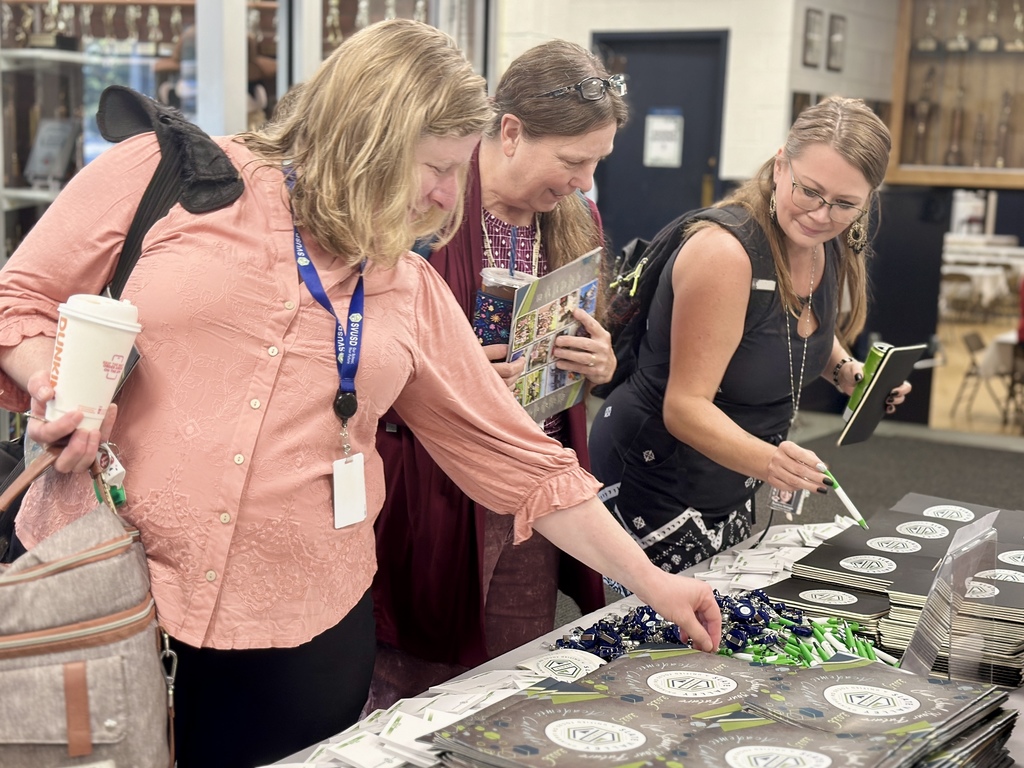 Three women stand at a table looking at Slate Valley Unified School District folders and pens. One woman in a pink shirt reaches for an item, while the others observe and smile.