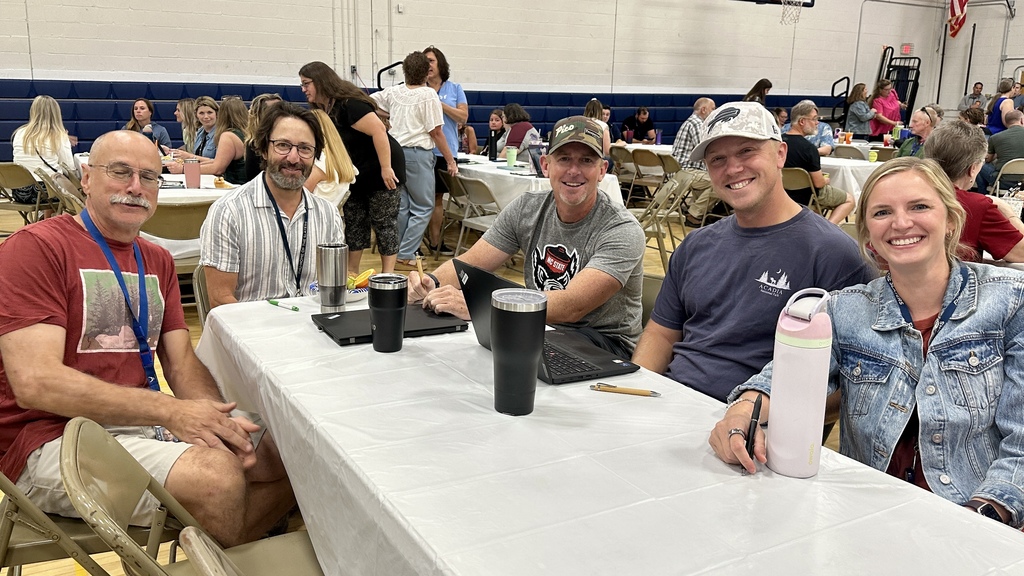 Five people sit together at a long table in a gymnasium during a school event. They are smiling at the camera, with laptops and tumblers in front of them. Other attendees can be seen seated at tables in the background.