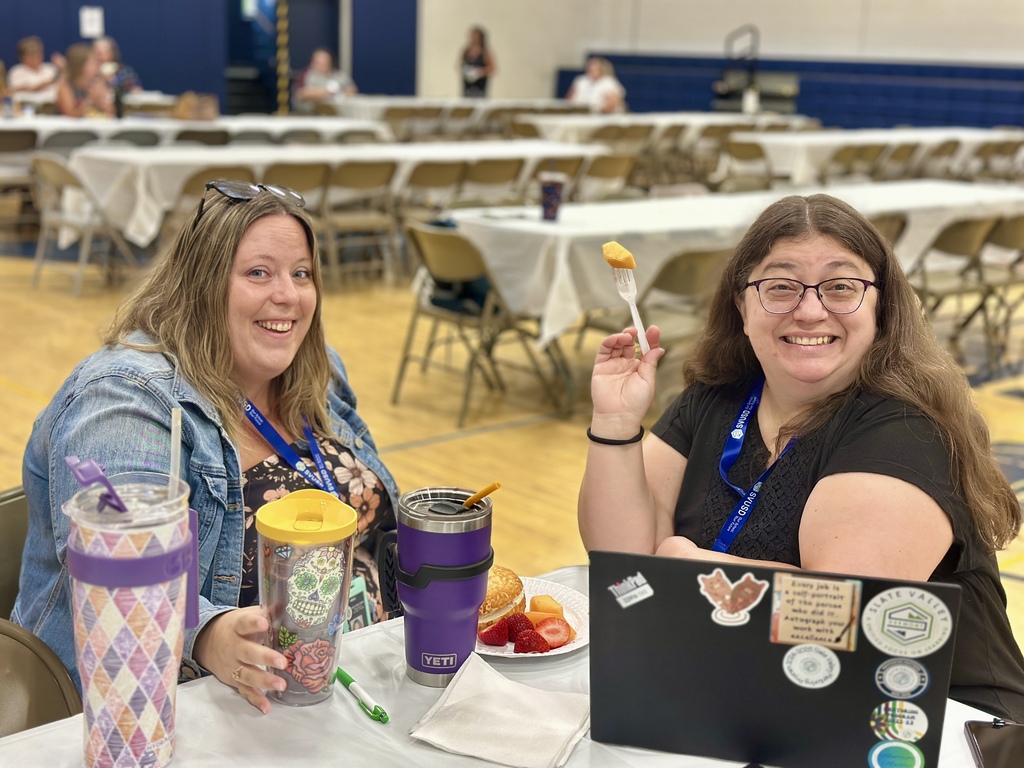 Two women sit at a table smiling, one holding a fork with fruit and the other with colorful tumblers. A laptop with stickers is on the table in front of them.