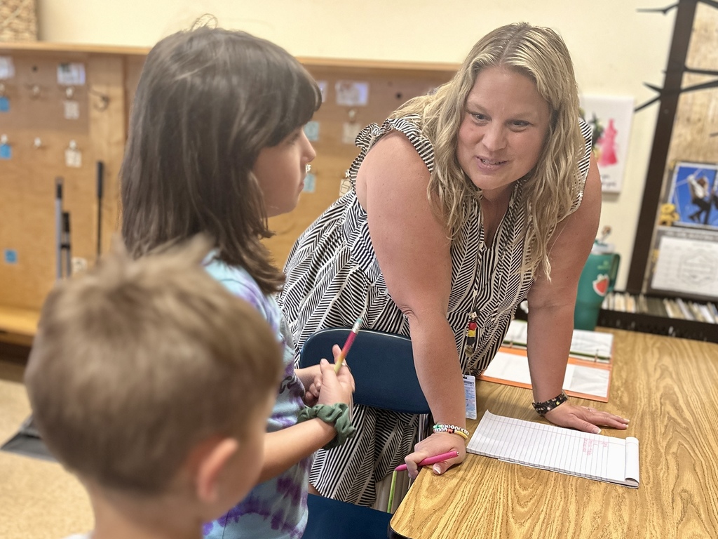 Teacher leans over a desk, smiling while talking with two young students.