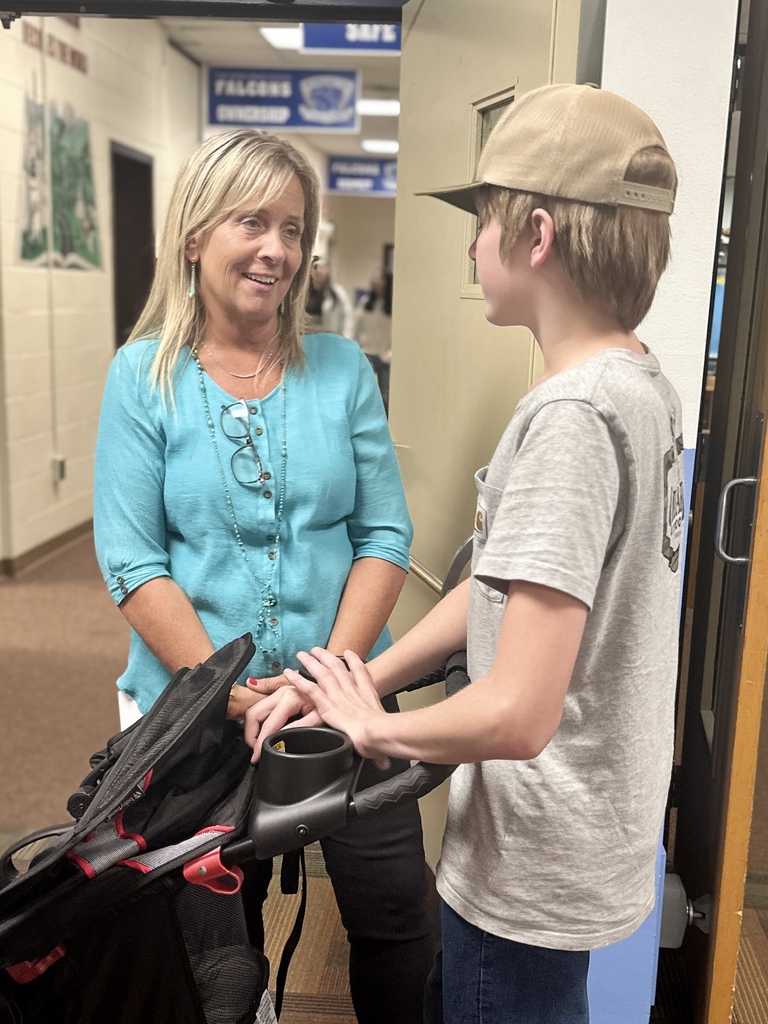 Teacher in a turquoise shirt talks with a student wearing a cap in a school hallway.