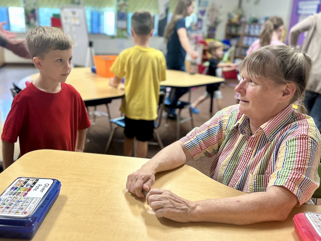 Teacher in a striped shirt sits at a table, smiling while talking with a young boy in a red shirt.