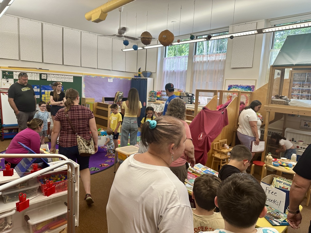Families and children gather in a busy classroom filled with books, toys, and activity areas.
