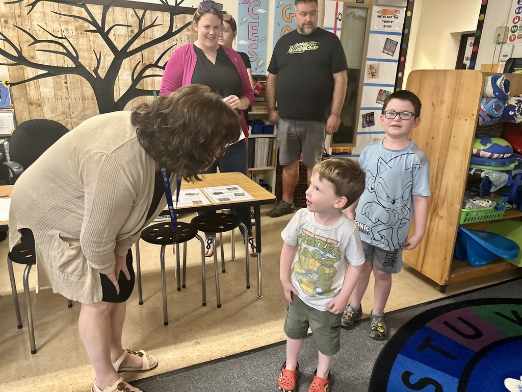Teacher bends down to greet two young boys in a classroom while families stand nearby.