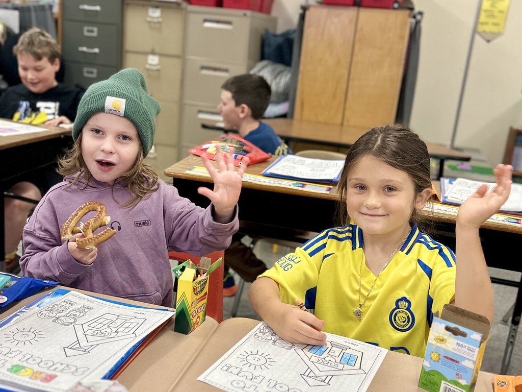 Two students wave while sitting at desks, one eating a pretzel, the other coloring.