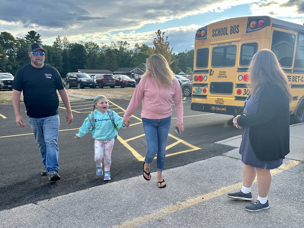 Family walks a young student toward the school, with a bus in the background.