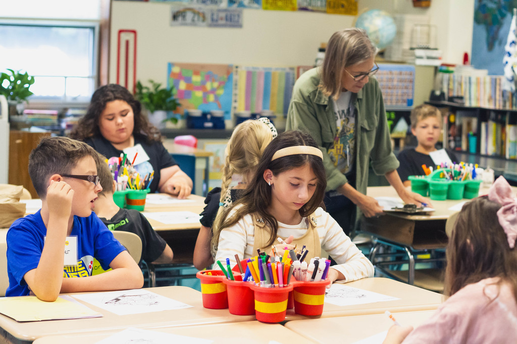 Students sit at desks coloring and writing, with a teacher assisting.
