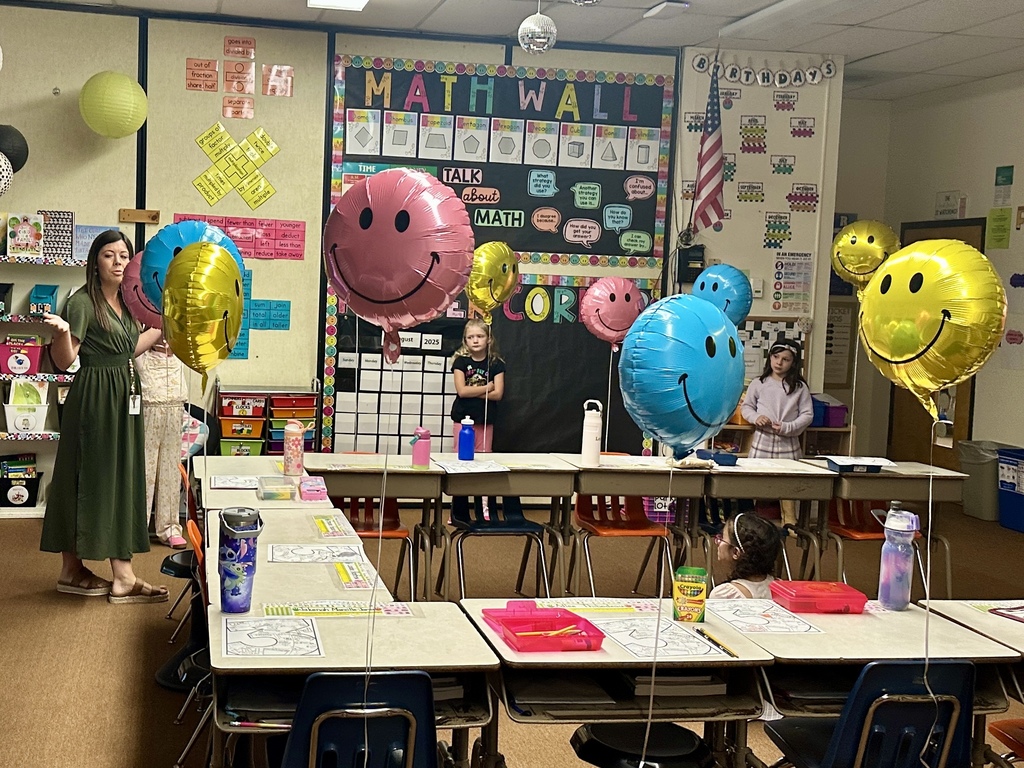 Classroom with desks decorated with worksheets and smiley face balloons; teacher addresses students.