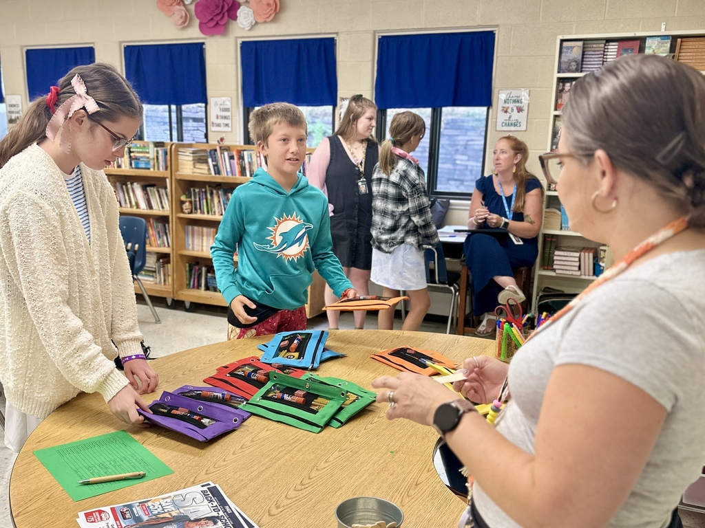 Students gather around a round table with colorful pencil pouches while a teacher assists in the background.