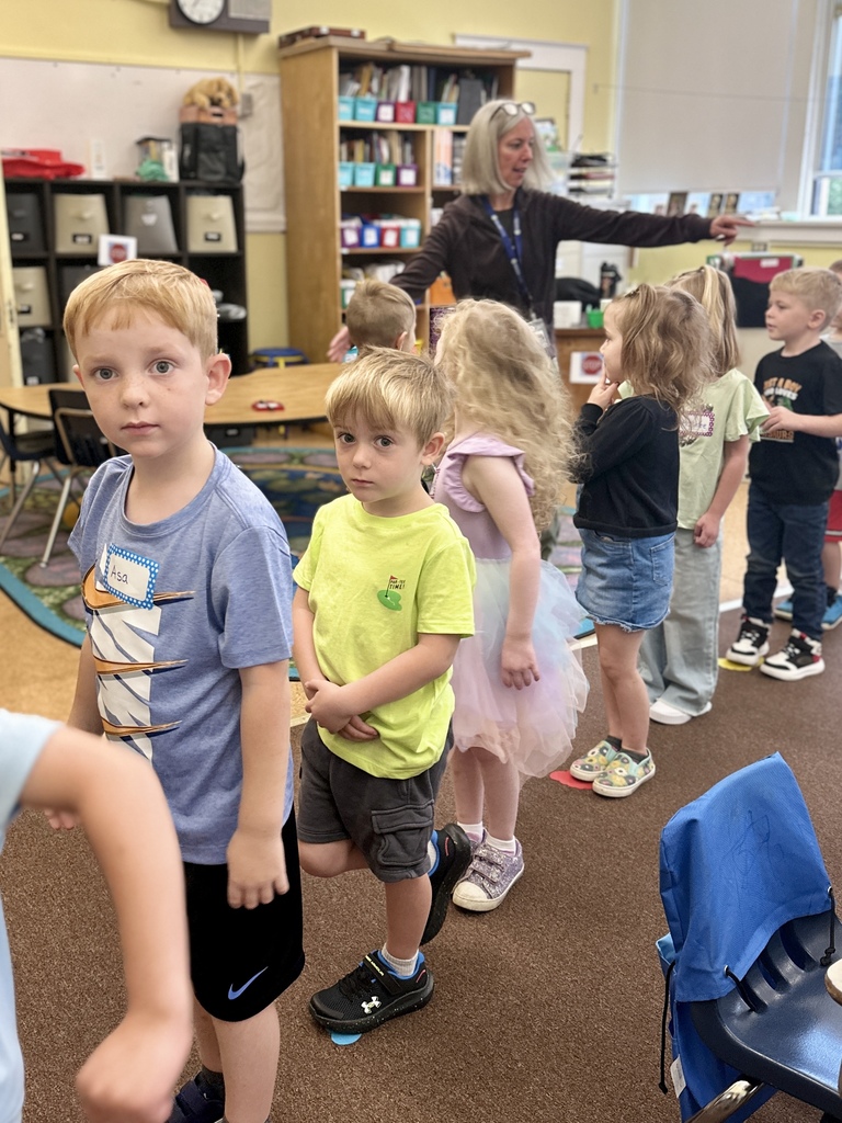 Young students line up inside a classroom as their teacher gives directions.