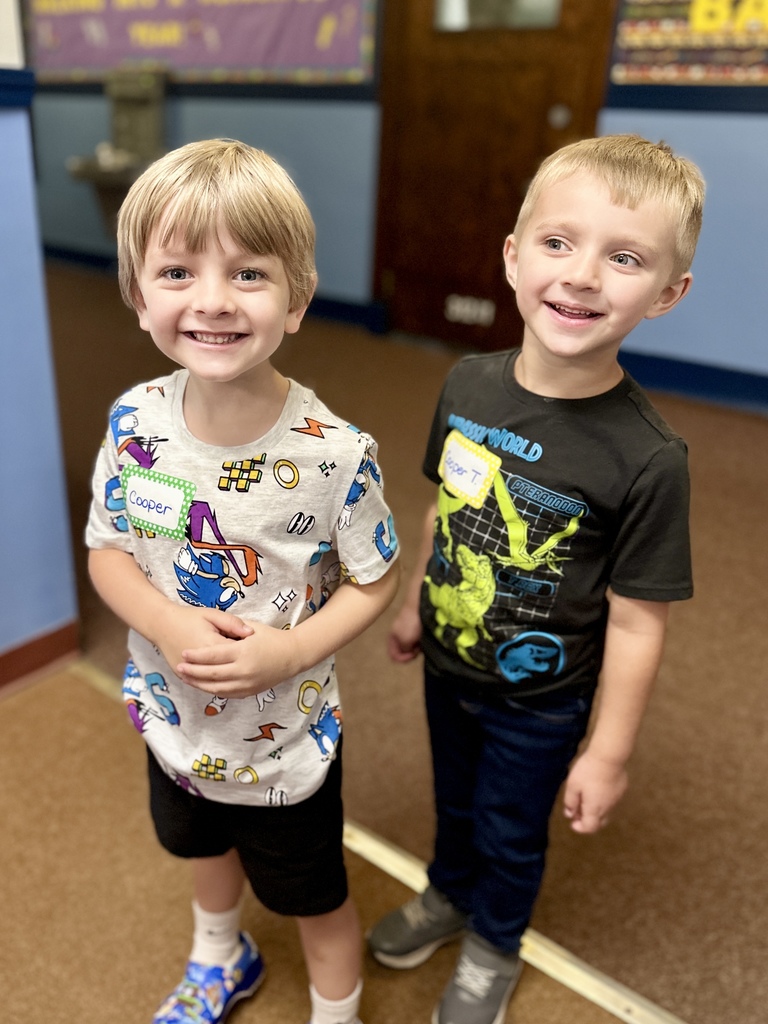 Two smiling boys wearing name tags stand together in a classroom hallway.