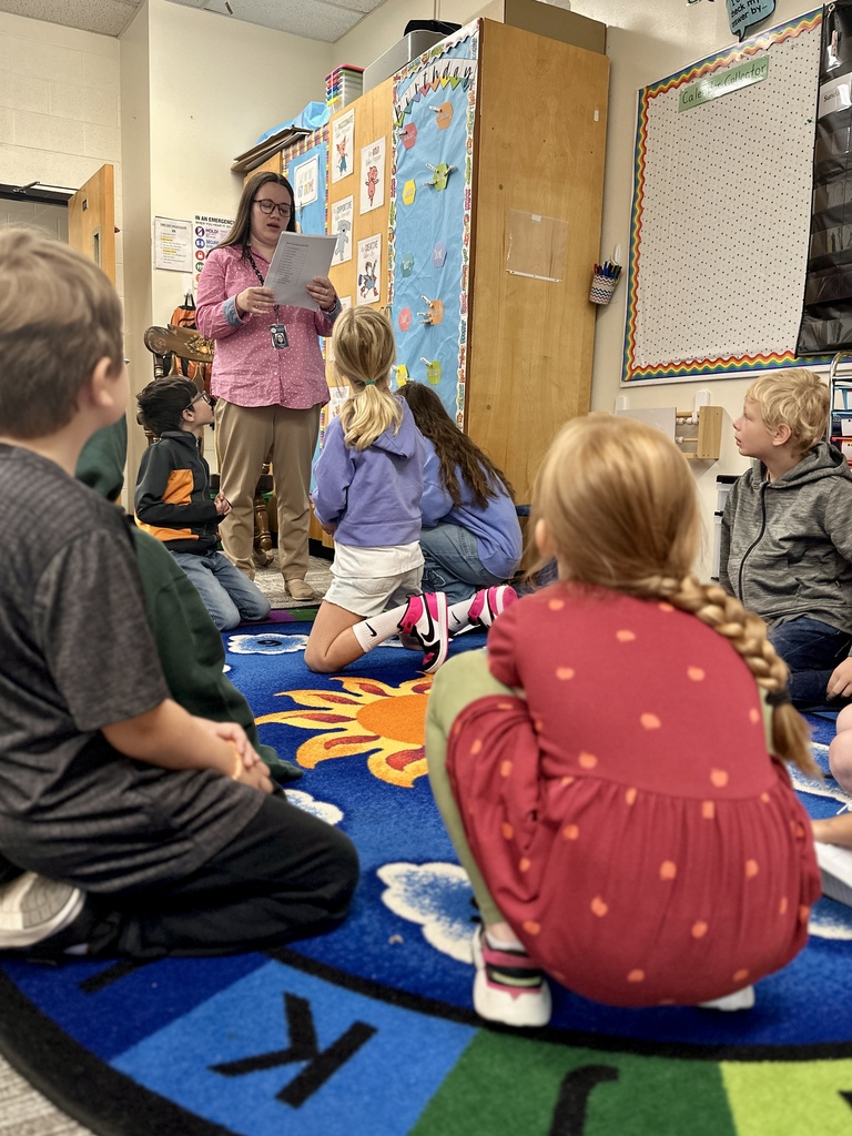 Teacher reads from a paper while students sit on a colorful rug, listening attentively.