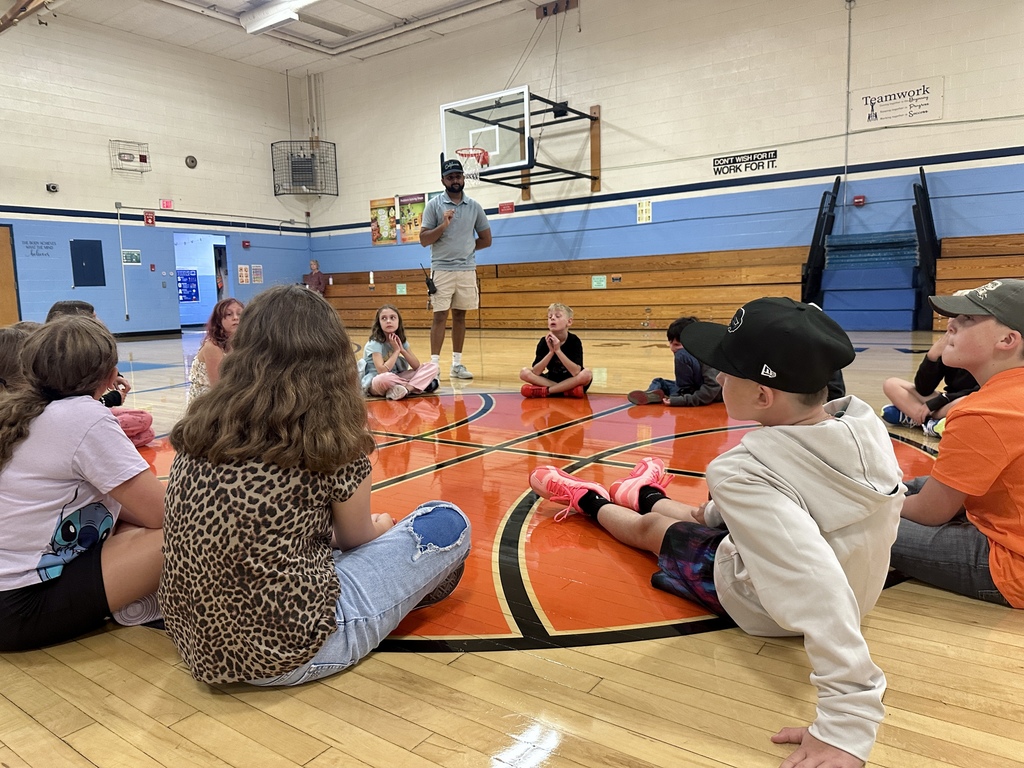 Students sit in a circle on the gym floor while a teacher gives instructions in the center.