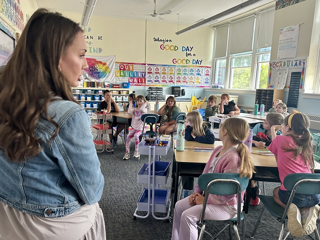 Teacher stands at the front of the classroom while students sit at desks listening attentively.
