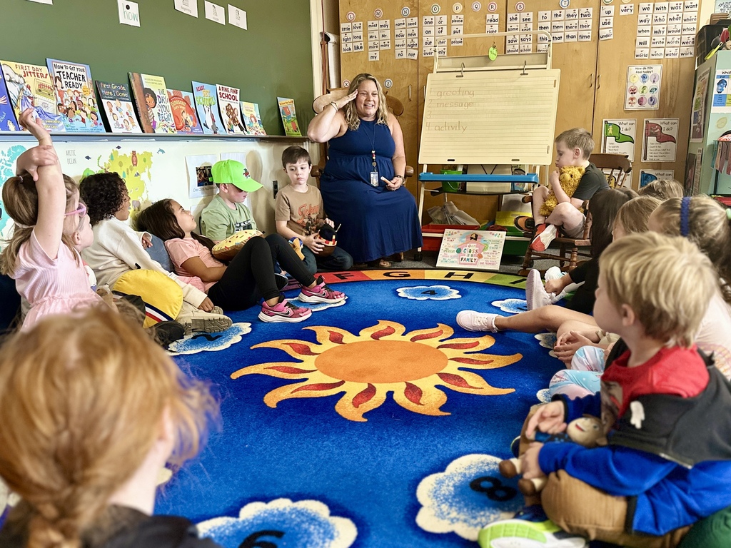 Teacher leads a morning circle; students sit on a rug decorated with a sun, raising hands to participate.