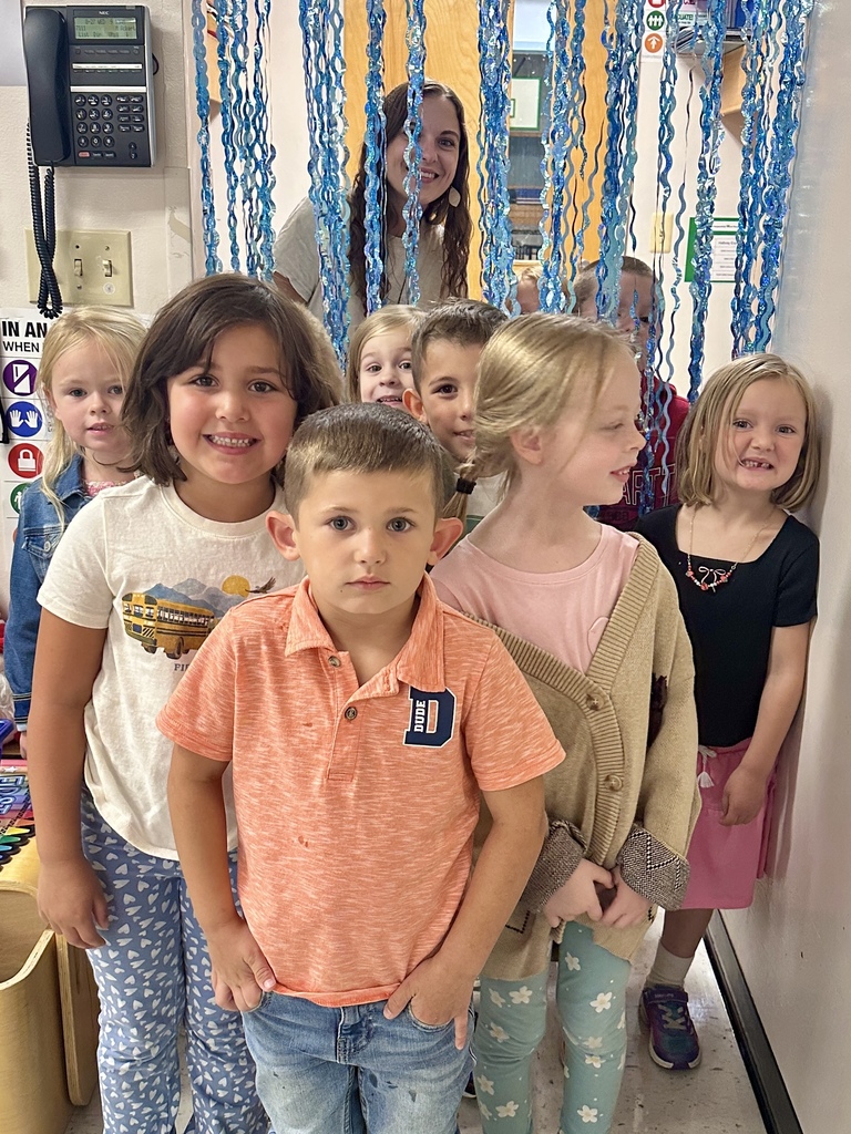 A group of young children stand together in a line, smiling, with blue hanging decorations behind them and a teacher peeking through in the background.