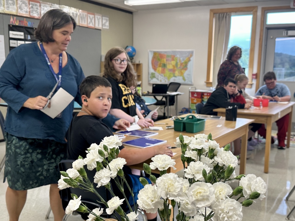 A teacher interacts with students in a classroom where children sit at tables working; a bouquet of white flowers is in the foreground.