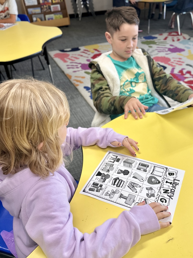 A young girl in a purple hoodie and a boy in a green shirt and camouflage jacket sit at a yellow table, looking at a "Library Bingo" worksheet.