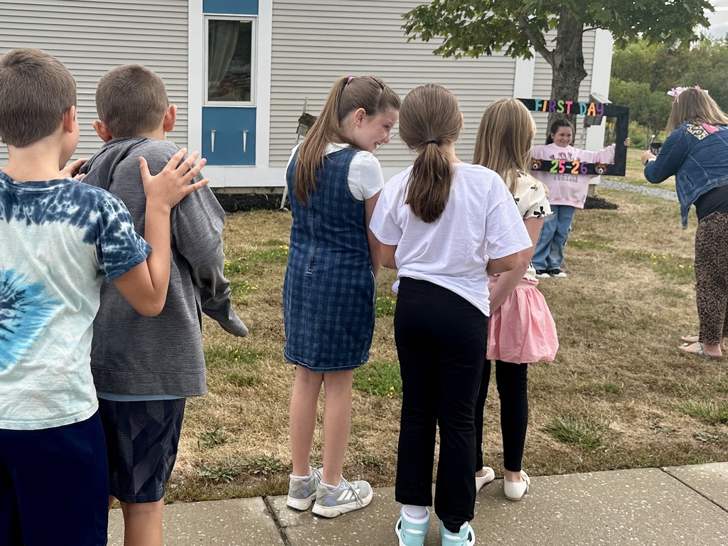 A group of students wait in line outdoors for their turn to pose with the “First Day 25–26” frame, while a classmate has their picture taken.