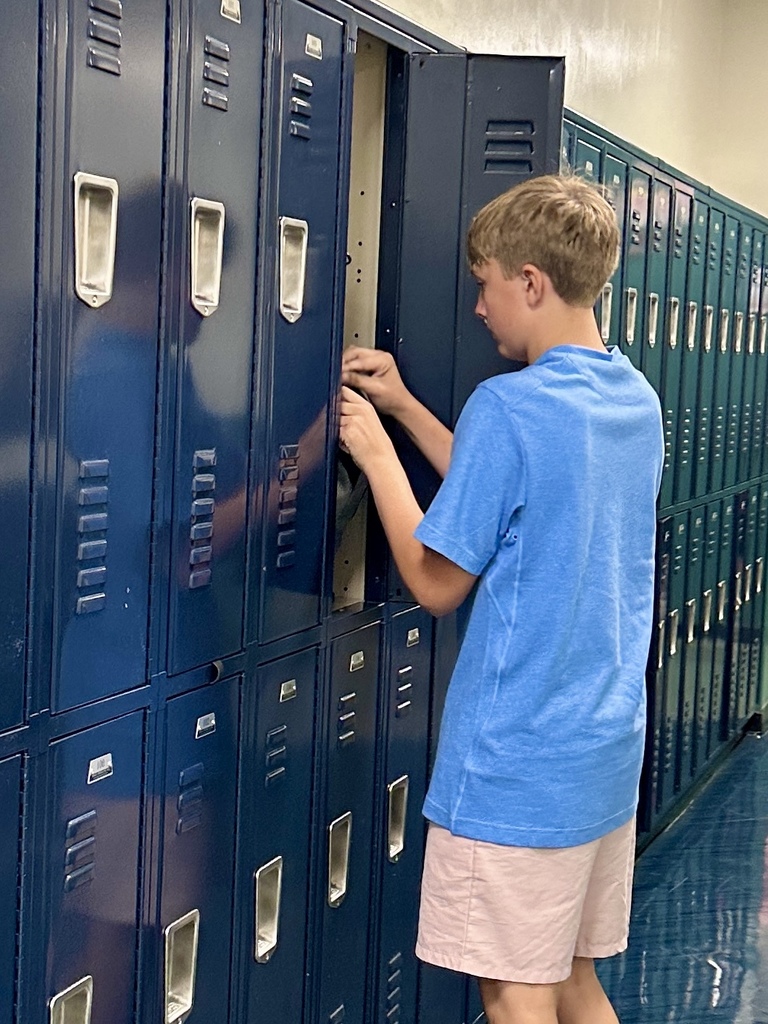 A boy in a blue shirt opens a tall, navy locker in a school hallway.