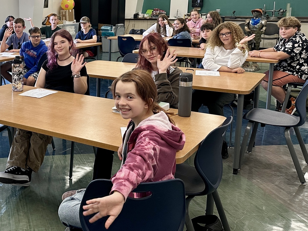 A group of middle school students sit at classroom tables and wave toward the camera, smiling.