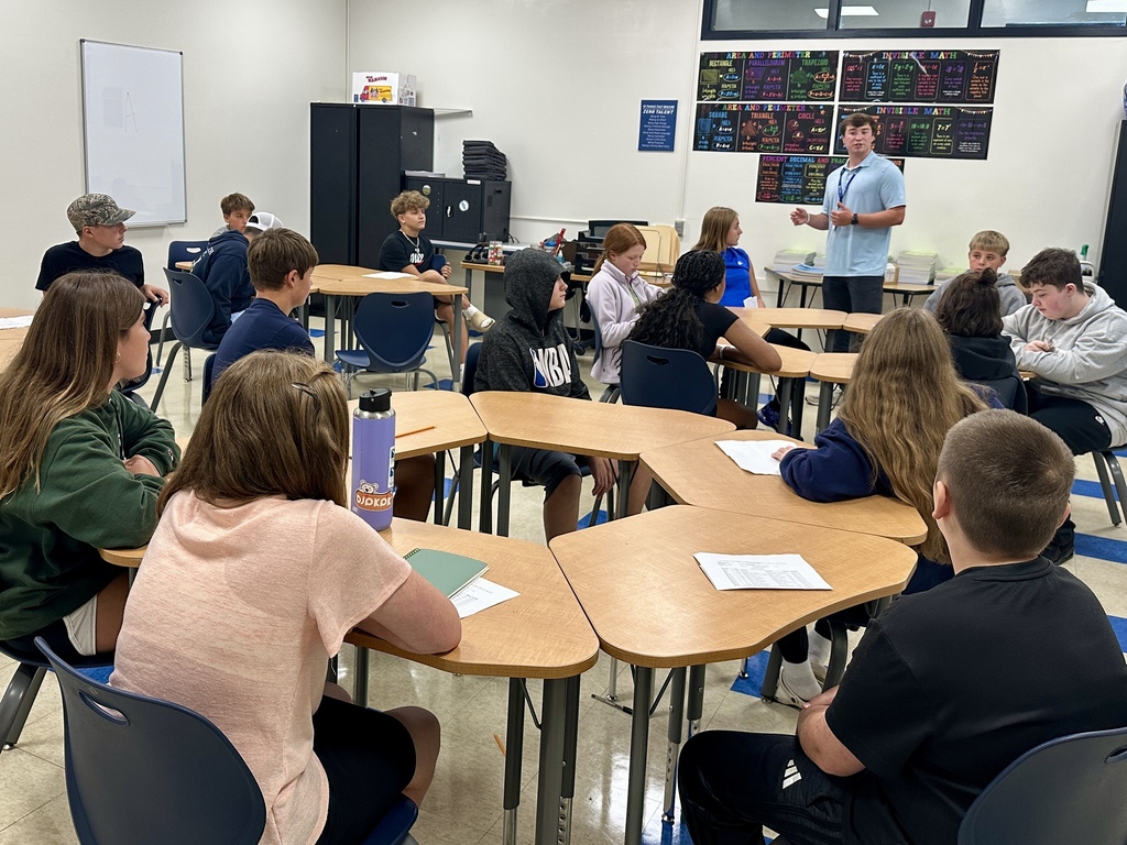 A male teacher stands and speaks to a group of middle school students seated at cluster desks, listening attentively.