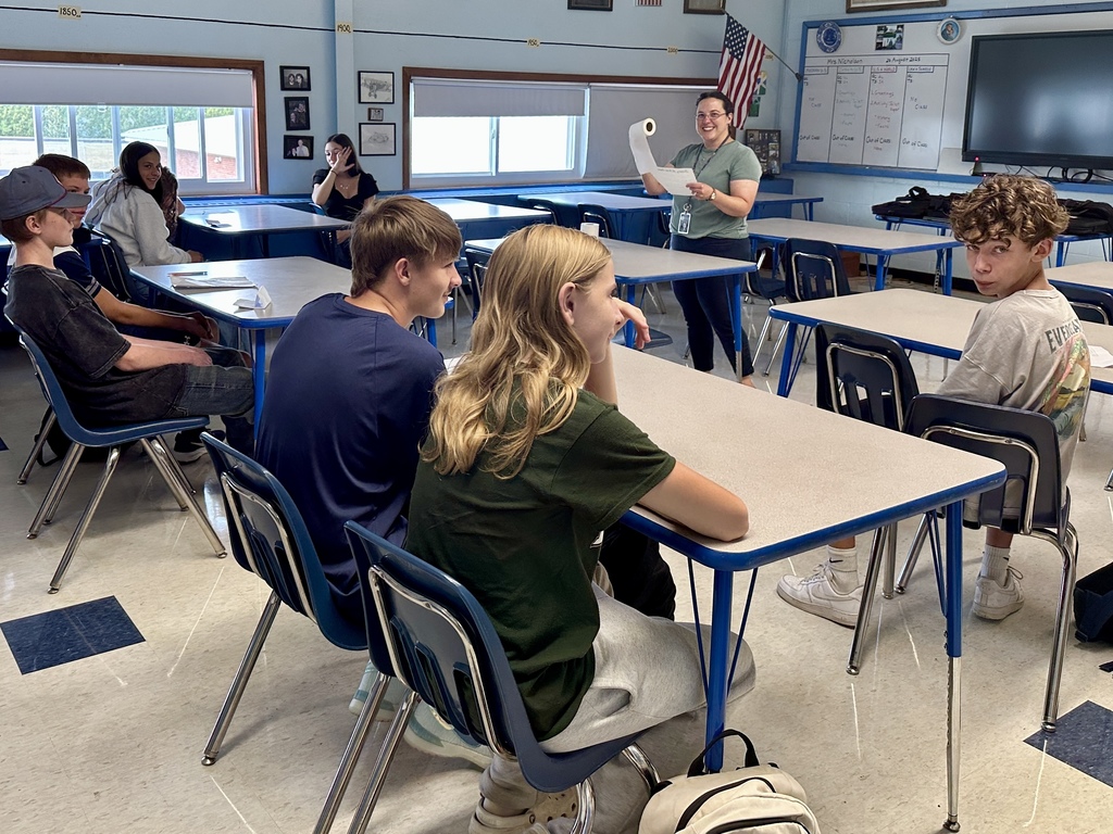 A female teacher stands at the front of a classroom holding a paper, while high school students sit at tables and listen.
