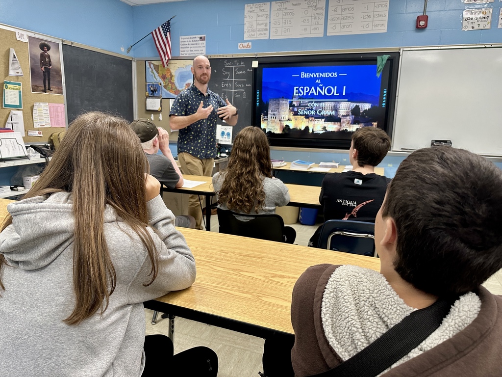 A Spanish teacher gestures while speaking to students, with a large screen behind him displaying “Bienvenidos al Español I.”
