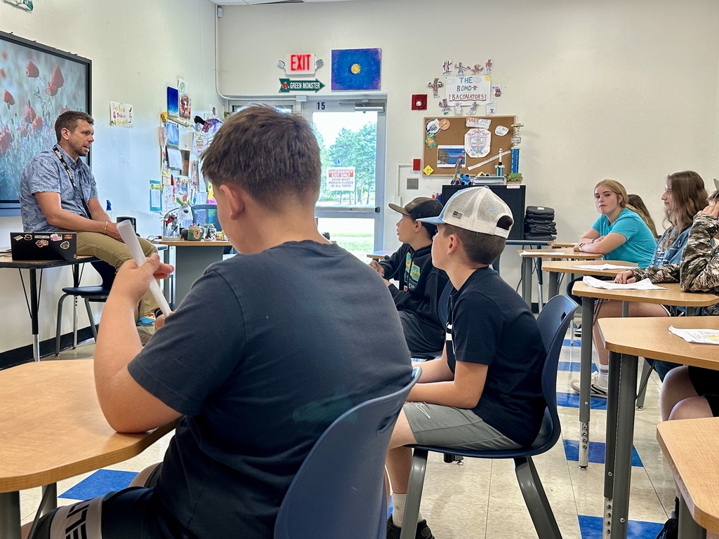 A male teacher sits at the front of the classroom, speaking to students sitting at desks. Posters and student work decorate the wall behind him.
