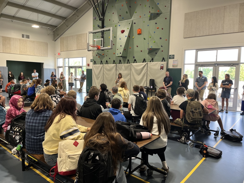 Middle school students sit at cafeteria tables in a gymnasium, listening to staff members lined up along the wall. A climbing wall and basketball hoop are visible in the background.