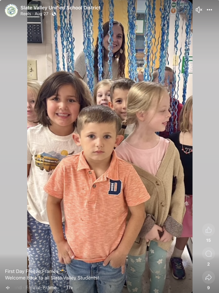 Screenshot showing young students posing for a photo with their teacher behind them peering through decorative blue streamers.