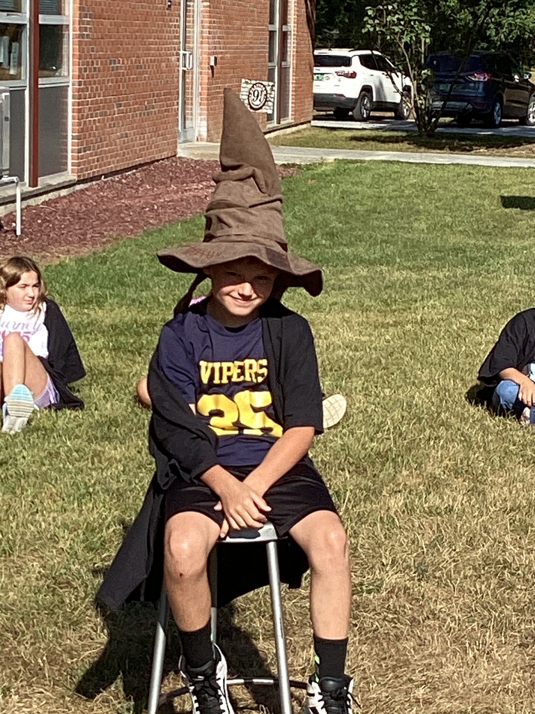 Student in a robe and Sorting Hat sitting on a stool outside on the grass.