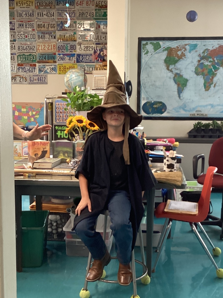Student in a robe wearing the Sorting Hat while sitting on a stool inside a classroom.