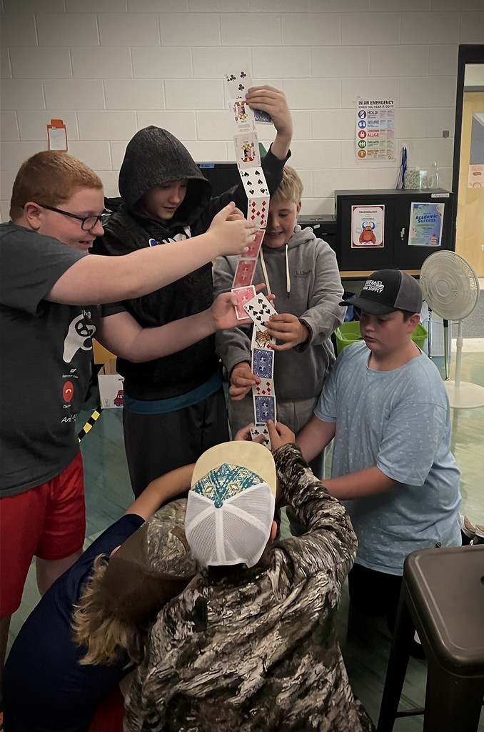 A group of boys work together to hold up a tall, vertical tower of playing cards.